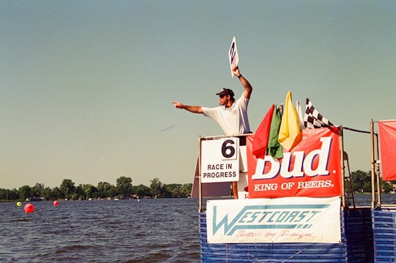 IJSBA - Finish line boat race marshal standing alone on race platform. Shown holding lap sign while pointing at racers and blowing whistle.