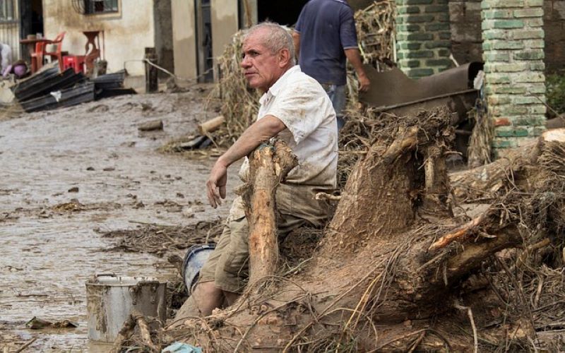 Post mass flood storm serge. Mud and debris covered streets. Exhausted man resting on tree stump. 