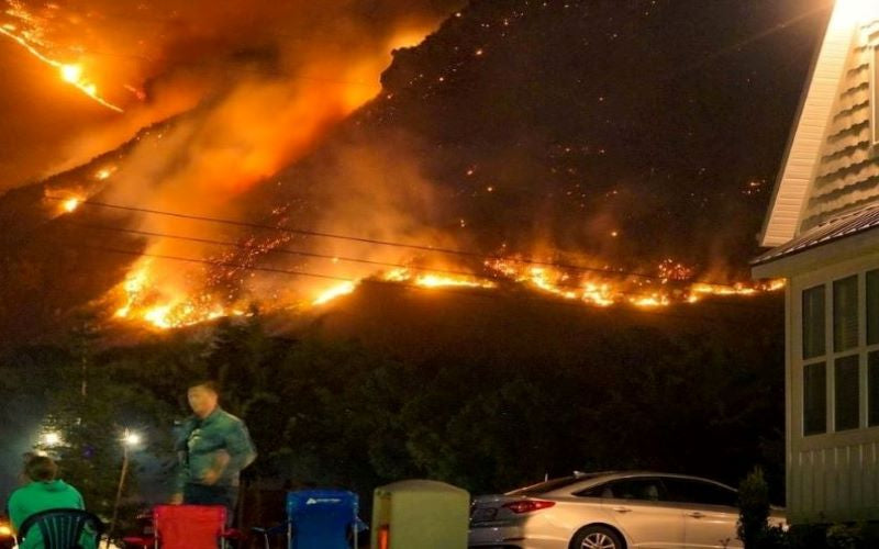 Mountainous Natural Disaster Wildfire at night approaching house, vehcles and people watching. 